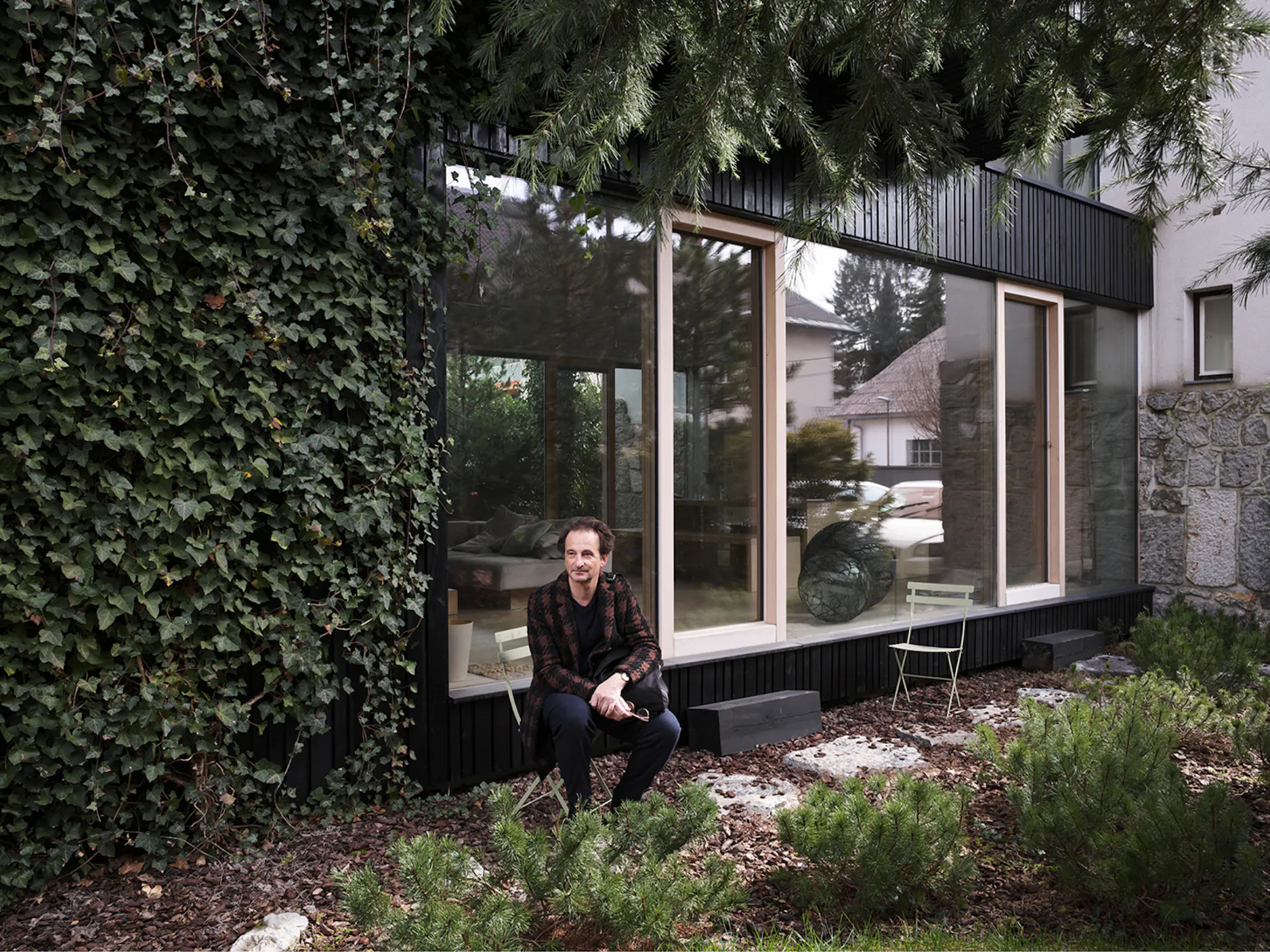 Moški sedi na stolu pred moderno hišo, okoli njega drevesa in grmičevje